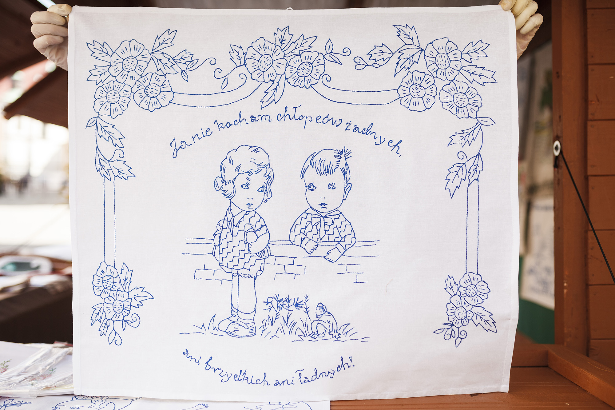 [Haft wykonany niebieską nicią na białej tkaninie, przedstawia chłopca z dziewczynką, dookoła nich napis:"ja nie kocham chłopców żadnych, ani brzydkich ani ładnych!"]