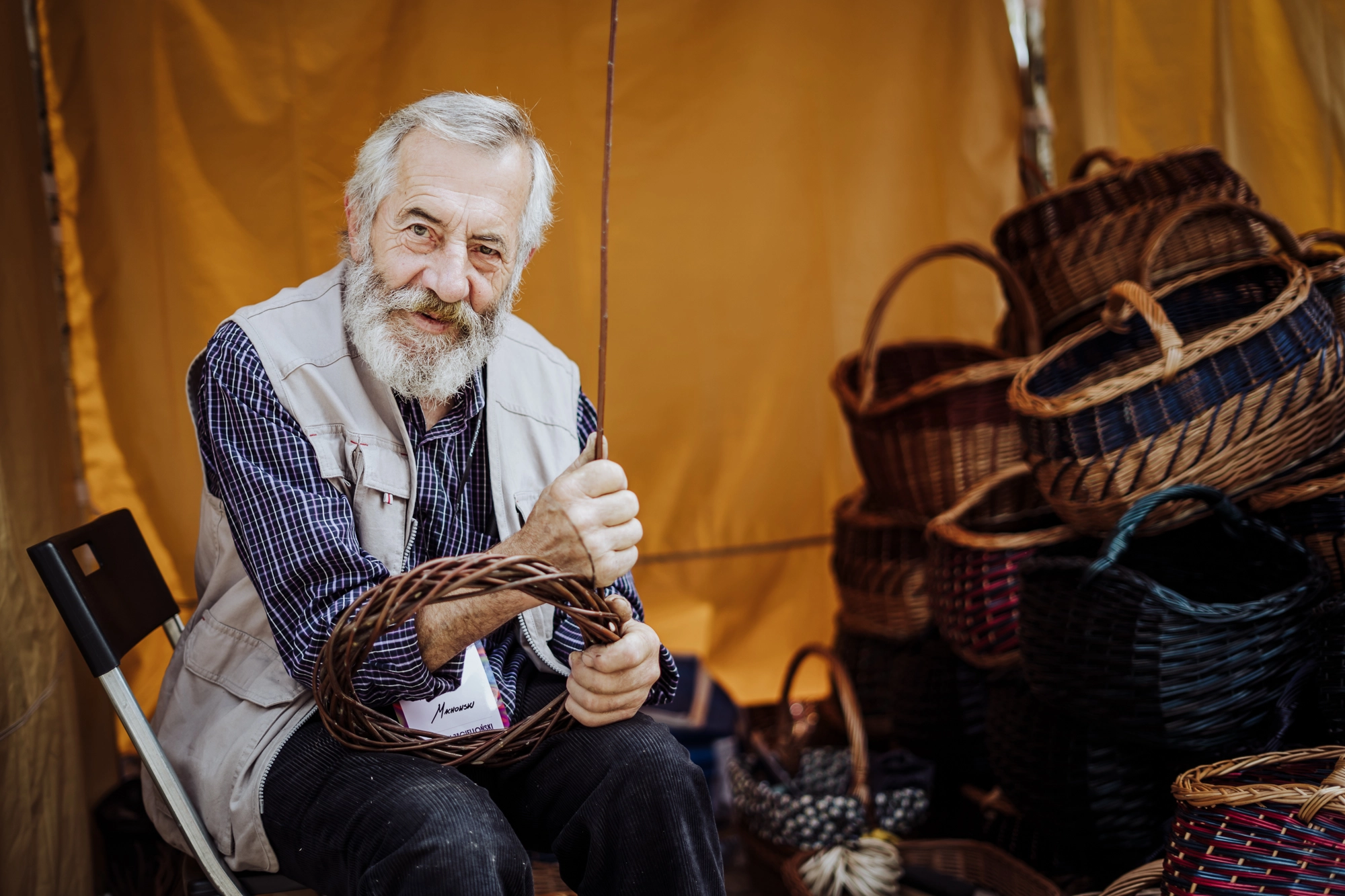 Portret twórcy. Starszy mężczyzna wyplata obręcz z wikliny. W tle kosze.