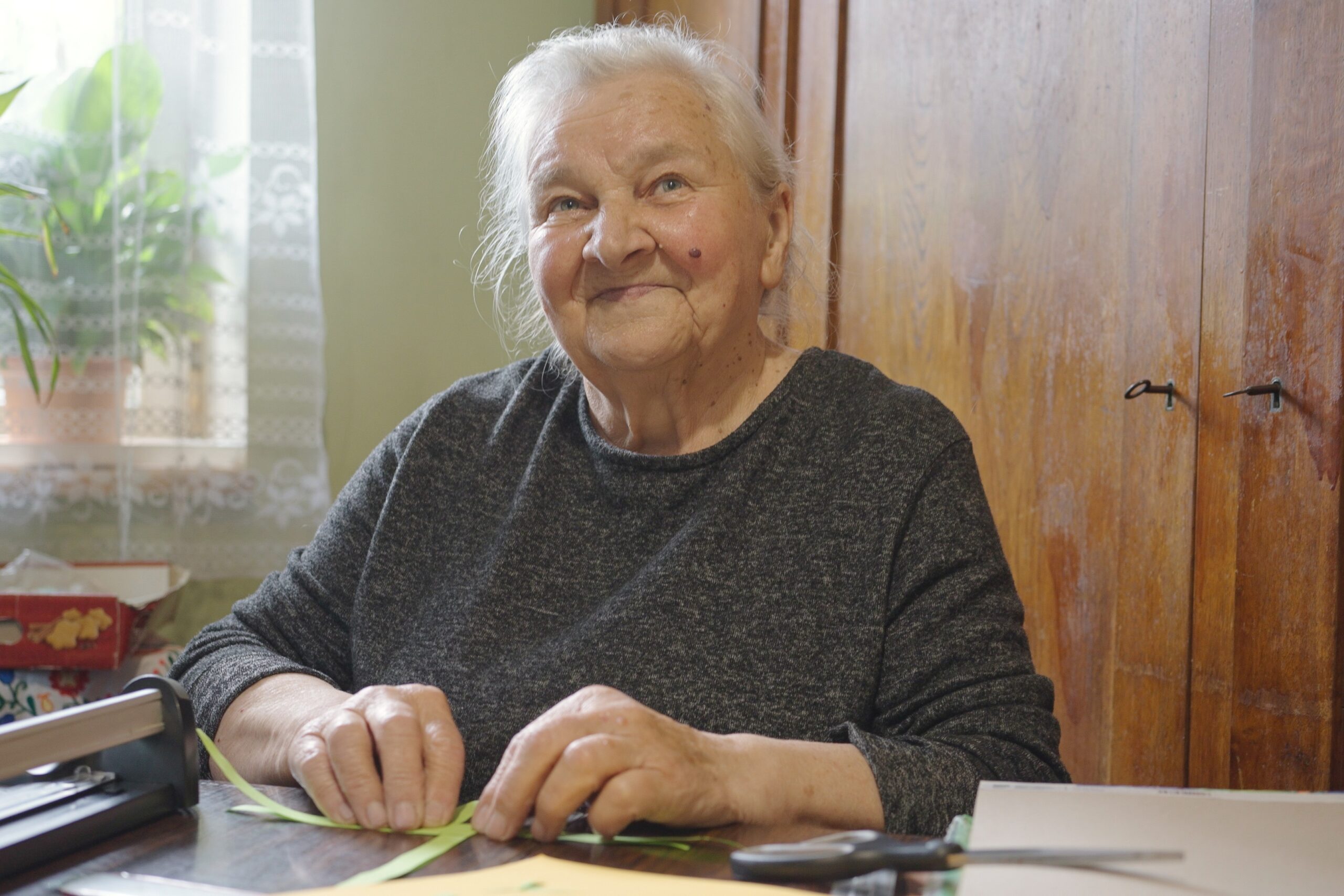 Portret twórczyni. Starsza kobieta siedzi przy stole. W dłoniach trzyma papier, z którego powstanie ozdoba choinkowa.