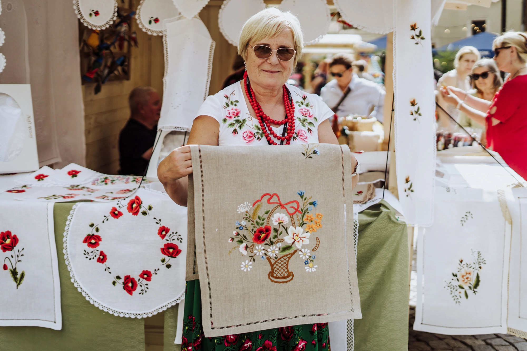 Portret twórczyni. Kobieta trzyma w rękach haftowaną makatkę. Wzór haftu to kosz pełen kwiatów.