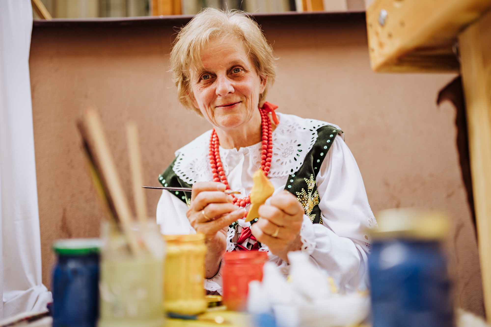 Portret kobiety, która jest w trakcie malowania drewnianego ptaka. Przed nią stoją kolorowe farby.