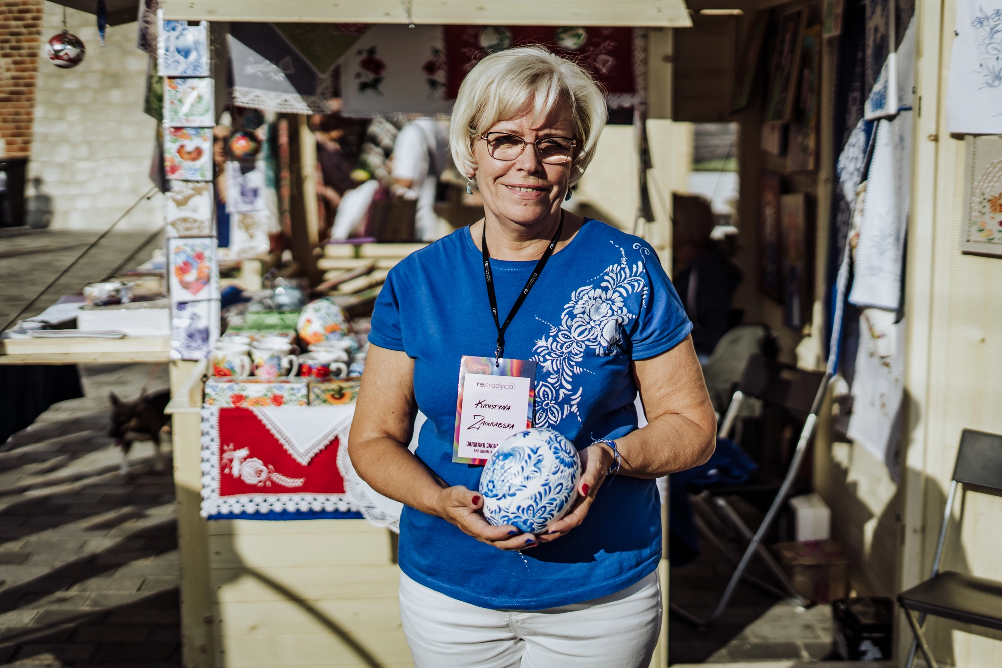 Portret twórczyni. Kobieta w niebieskiej koszulce ozdobionej białym, malowanym wzorem kwiatowym. W rękach trzyma białą pisankę ozdobioną niebieskimi wzorami. To wzory kujawskie, znane na przykład z fajansu włocławskiego.