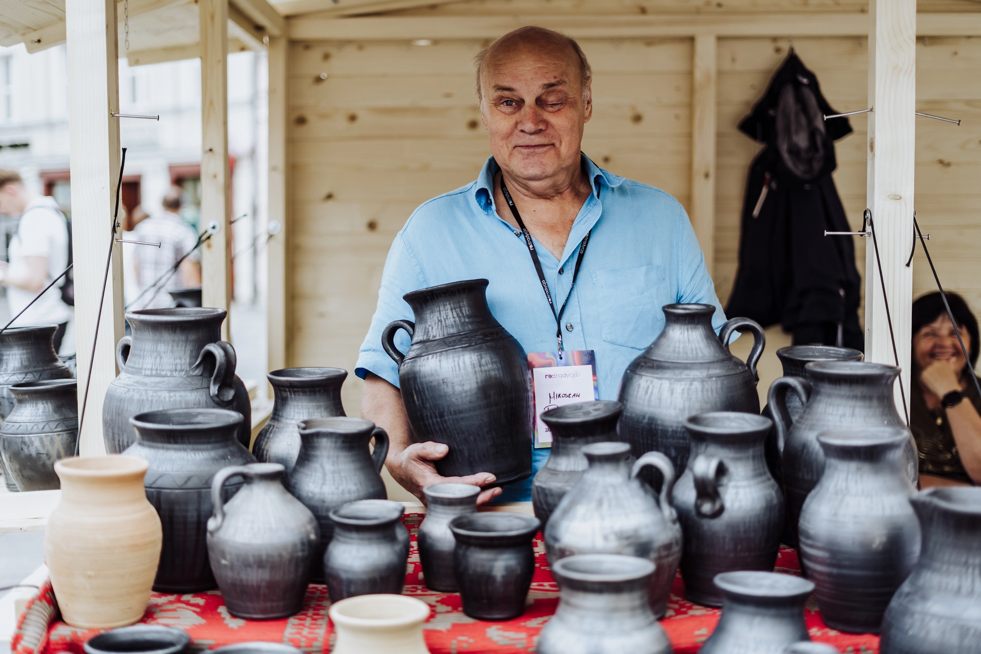 Portret twórcy. Mężczyzna stoi przy stoisku pełnym glinianych naczyń. Ceramika jest w kolorze czarnym i ma bardzo delikatne wzory.