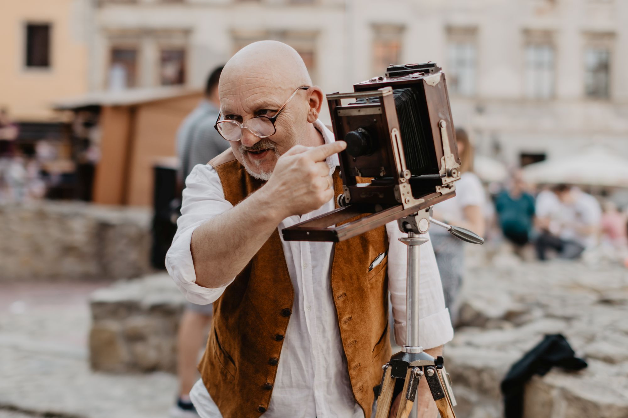 Roman Krawczenko, fotograf, wskazuje palcem na obiektyw zabytkowego aparatu fotograficznego, którym wykonuje zdjęcia. Starszy mężczyzna ma na sobie duże okulary, białą koszulę i miodową kamizelkę.
