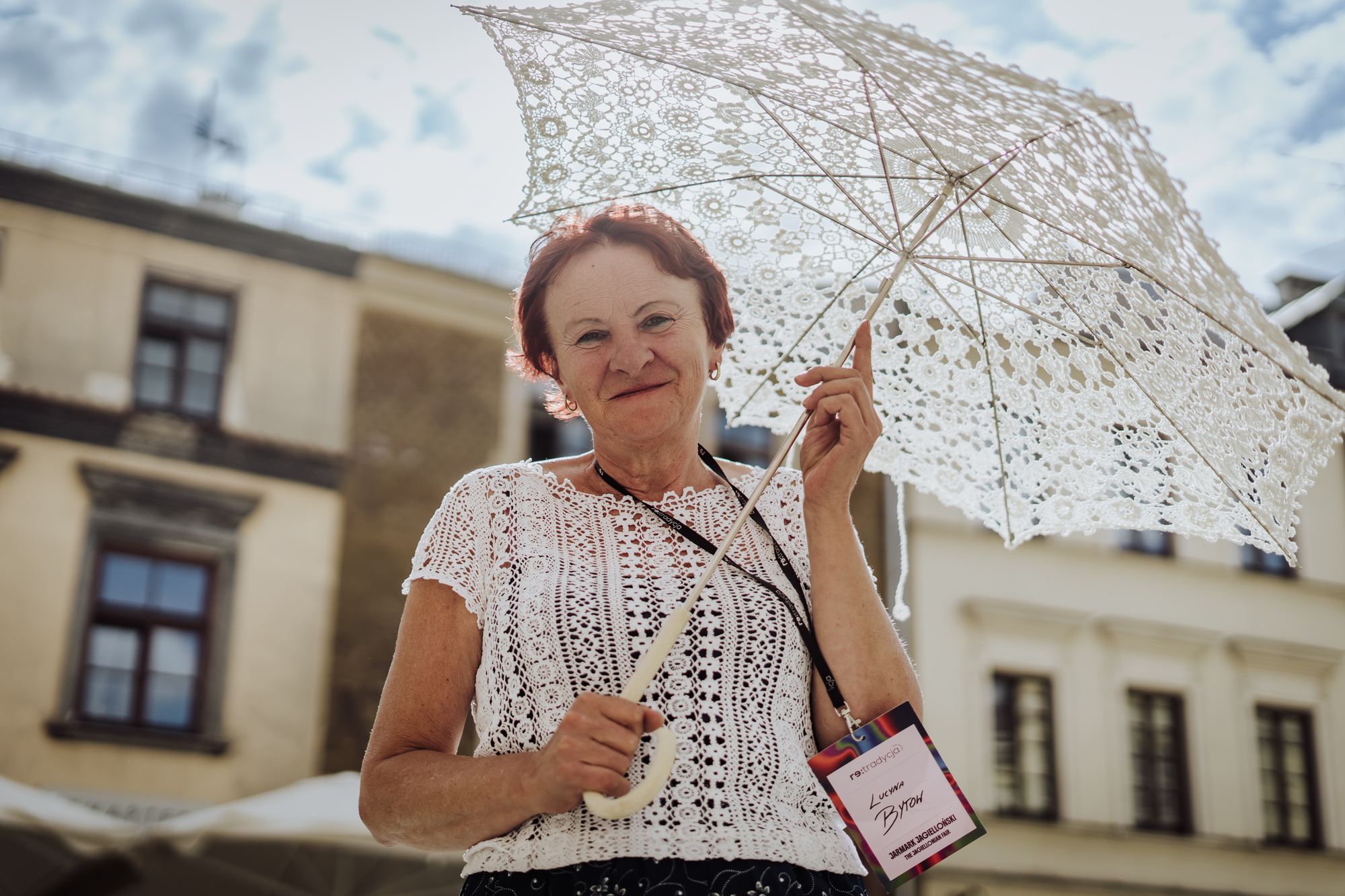 Portret twórczyni. Uśmiechnięta kobieta trzyma w rękach białą, koronkową parasolkę. W tle staromiejskie kamienice.