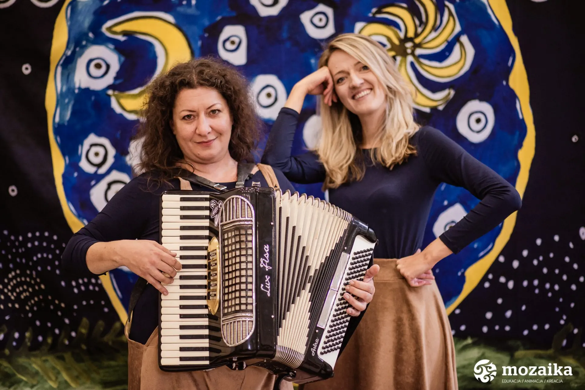 Kobieta gra na akordeonie. Obok stoi jej koleżanka.