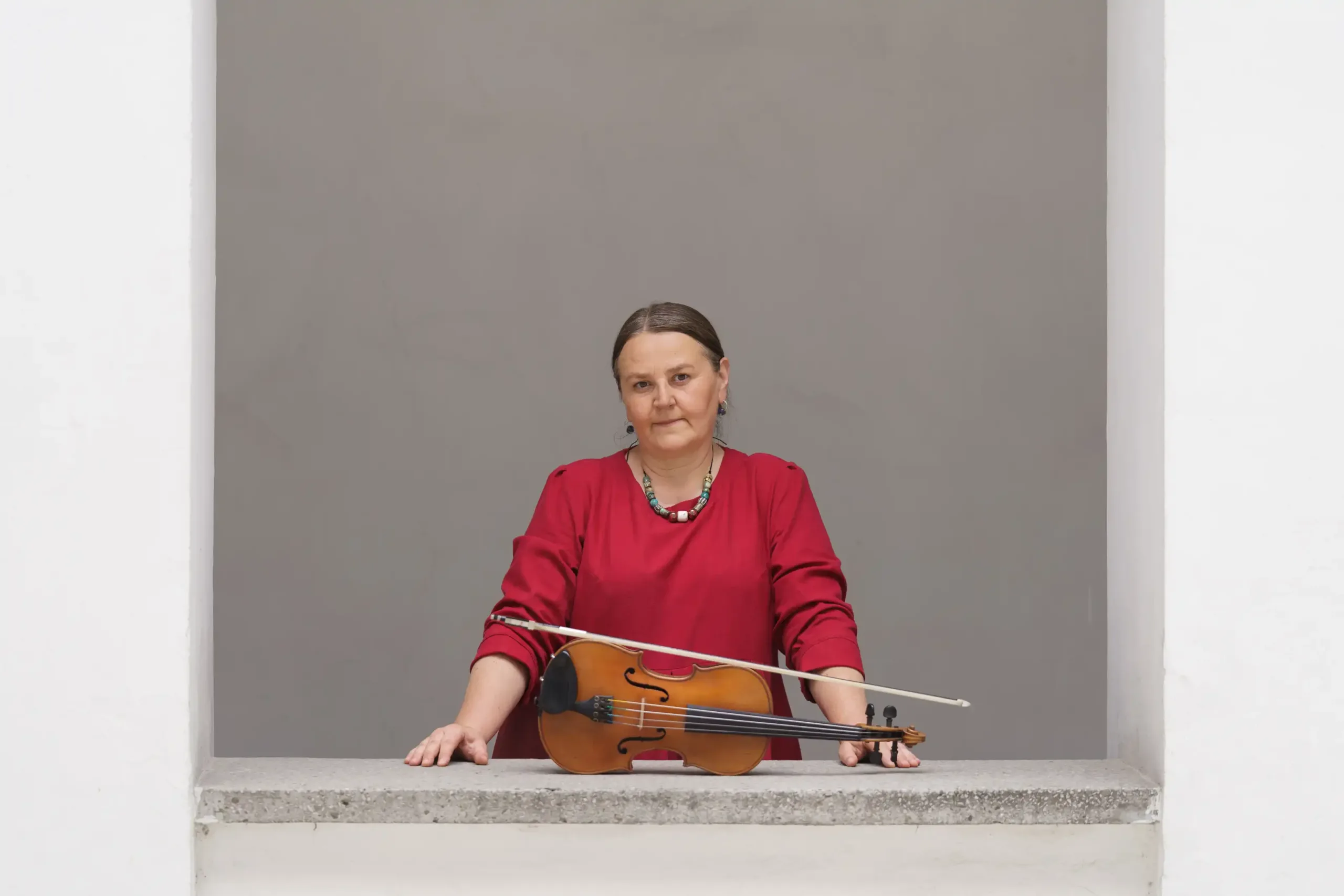 Portret artystki. Kobieta w czerwonej sukience ma przed sobą skrzypce i smyczek. Patrzy prosto w obiektyw.