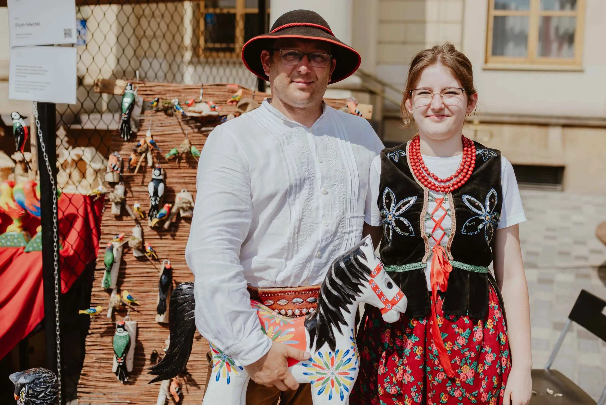 Twórca ludowy z córką. Oboje w strojach ludowych. Mężczyzna trzyma w dłoniach drewnianego konika.