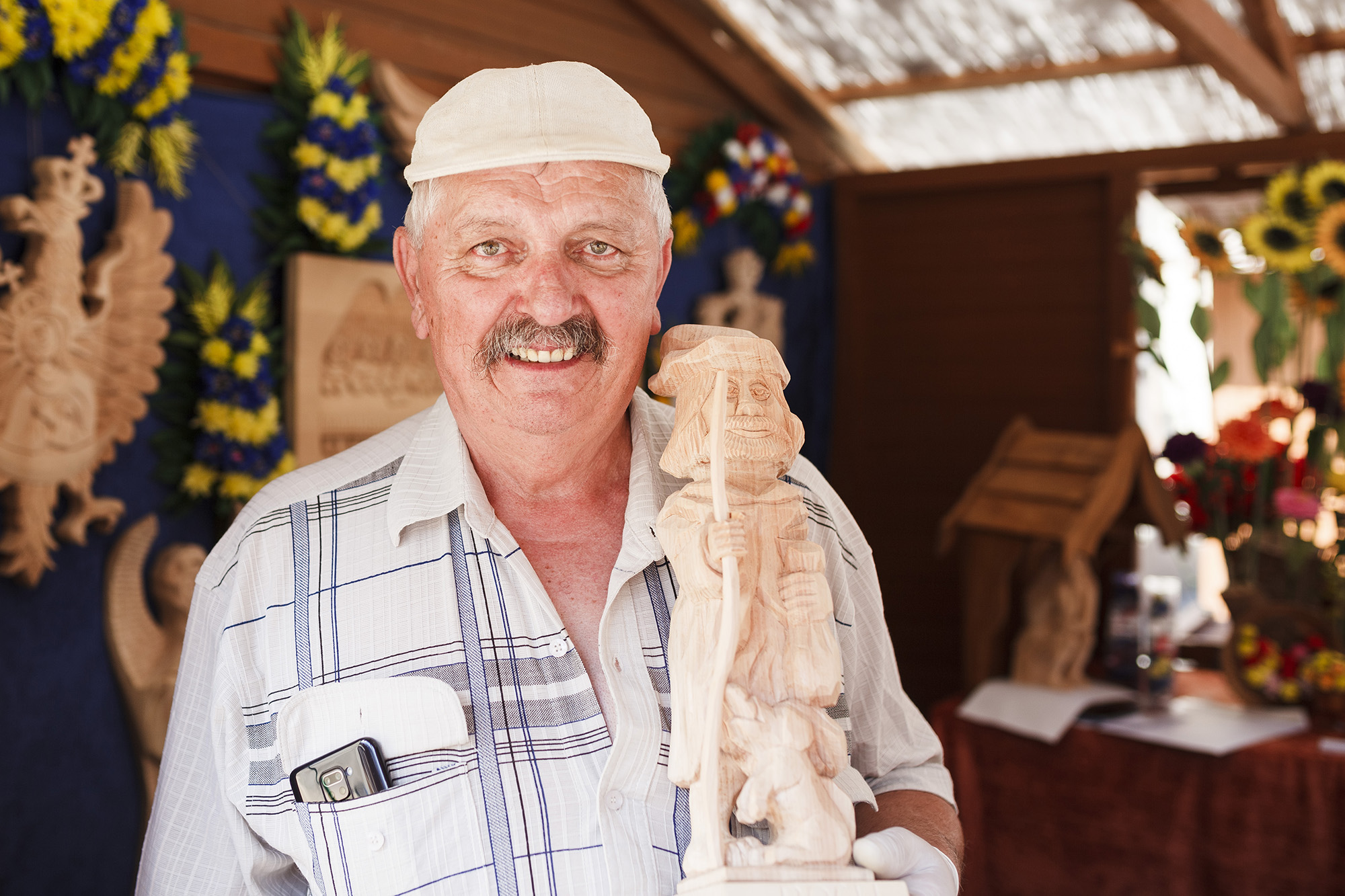 A smiling man in a checkered shirt is holding a wooden sculpture 