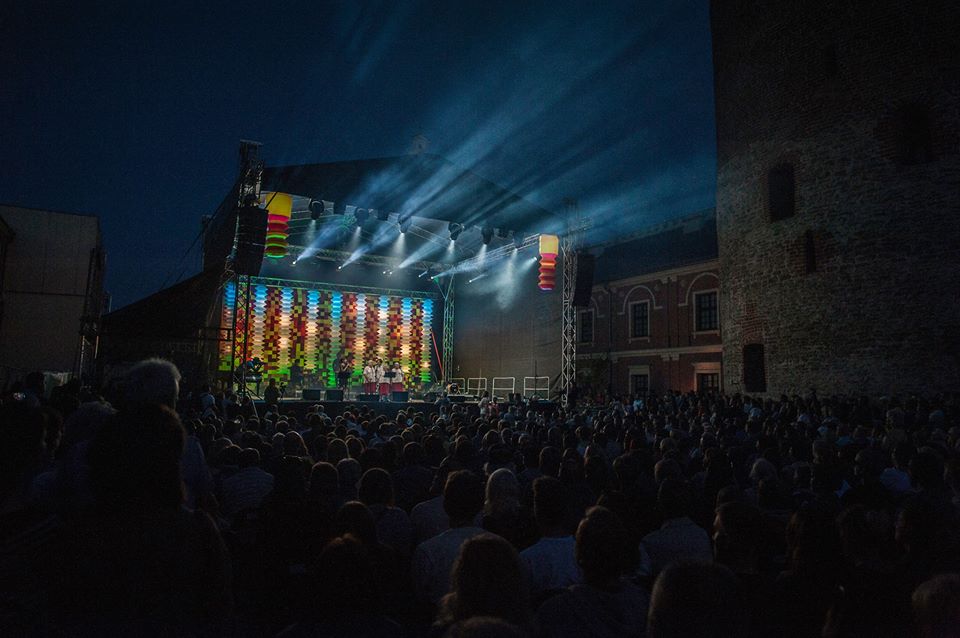 Noc, Dziedziniec Zamku Lubelskiego. Z dużej sceny wypływają w przestrzeń nad publicznością światła jasnych reflektorów. Scena wypełniona jest kolorową, migoczącą scenografią.