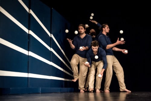 [W tle ciemnego kadru stoi skupiona blisko siebie grupa czterech mężczyzn żonglujących białymi piłeczkami. Obok nich, na granatowej ścianie biegną cztery poziome, białe linie.]