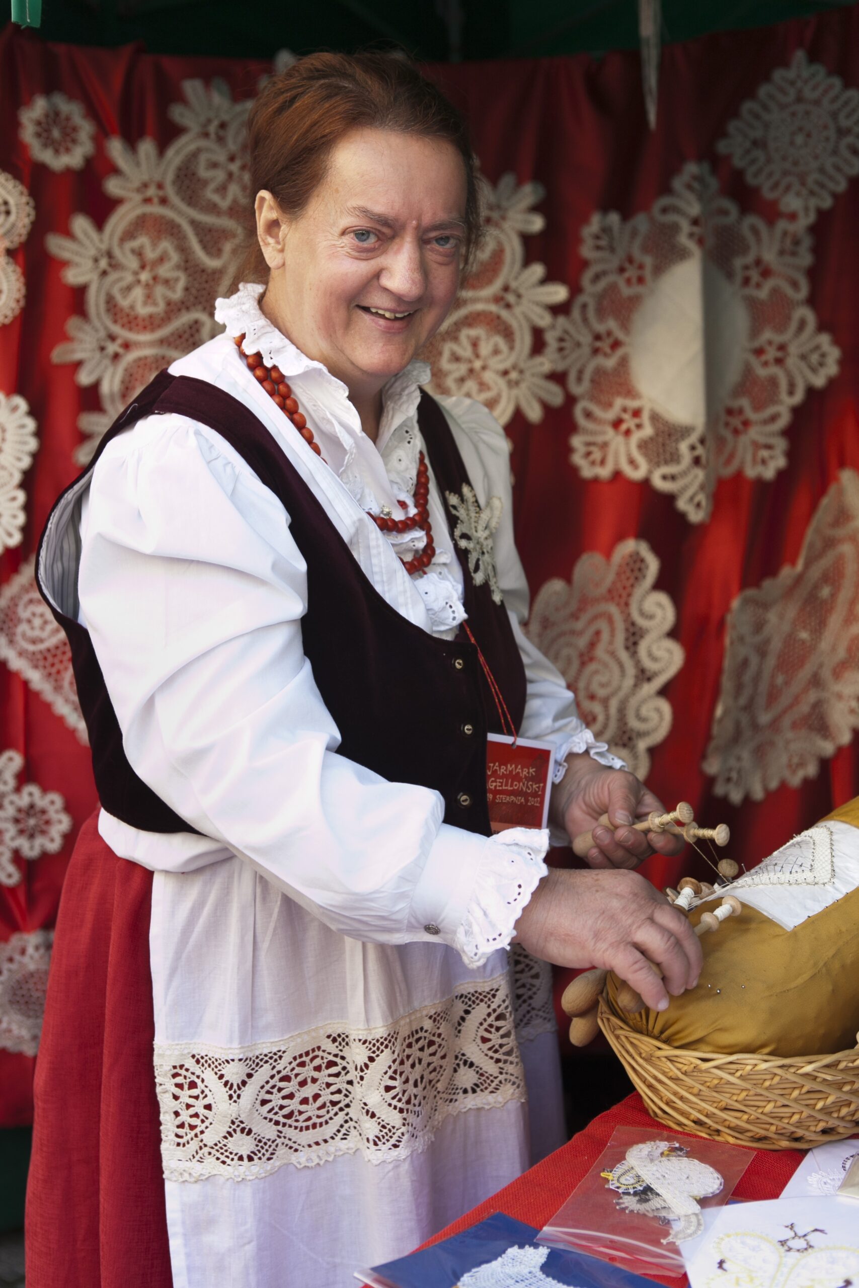 [Kobieta stoi na swoim stoisku z tradycyjnym rękodziełem.]