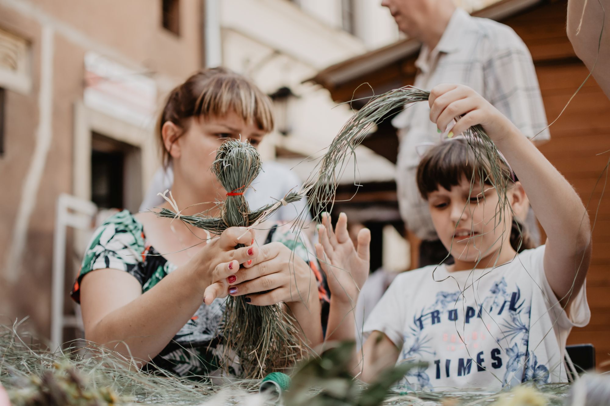 Mama i córka podczas warsztatów. Kobiety wykonują wspólnie laleczkę z siana. Matka trzyma w dłoniach częściowo ukształtowaną lalkę, córka dokłada do niej kolejne elementy.