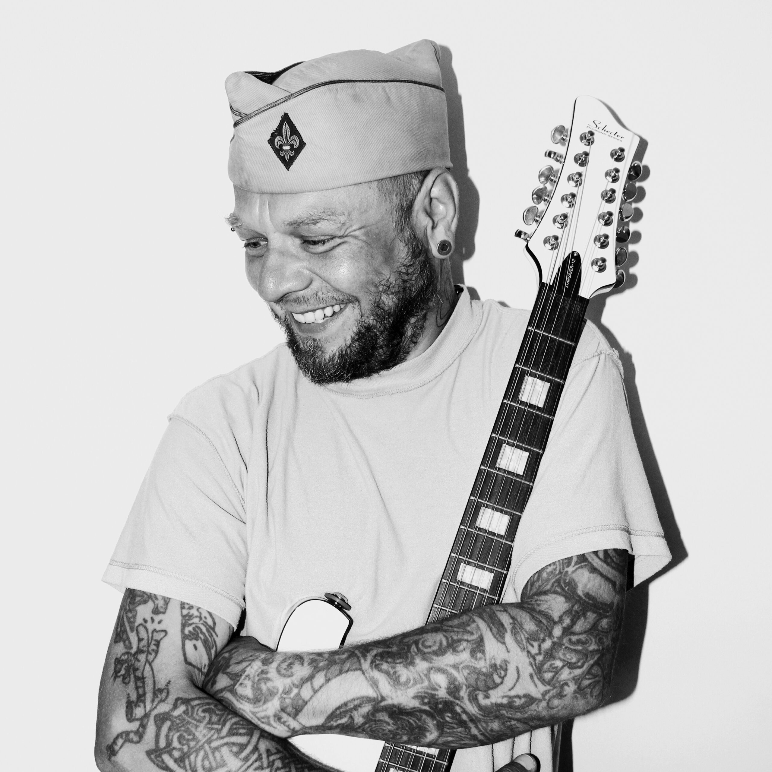 Czarno-biały portret Tomasza "Lipy" Lipnickiego, muzyka. Mężczyzna ma na przedramionach tatuaże, a na głowie czapkę, furażerkę. Rękami obejmuje gitarę elektryczną.