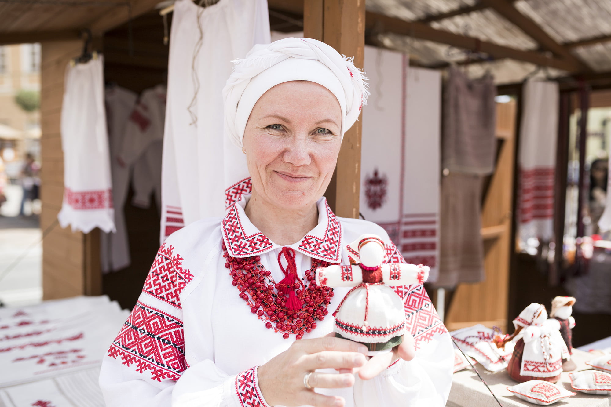 [Zbliżenie. Uśmiechnięta kobieta w stroju ludowym prezentuje swoje wyroby związane z haftem tradycyjnym.]
