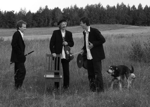 Czarno-białe zdjęcie. Na łące pod lasem stoi trzech mężczyzn. Ubrani w za duże, czarne marynarki, białe koszule i ciemne spodnie, rozmawiają ze sobą, trzymając instrumenty - klarnet, skrzypce i baraban. Obok nich kręcą się dwa psy.