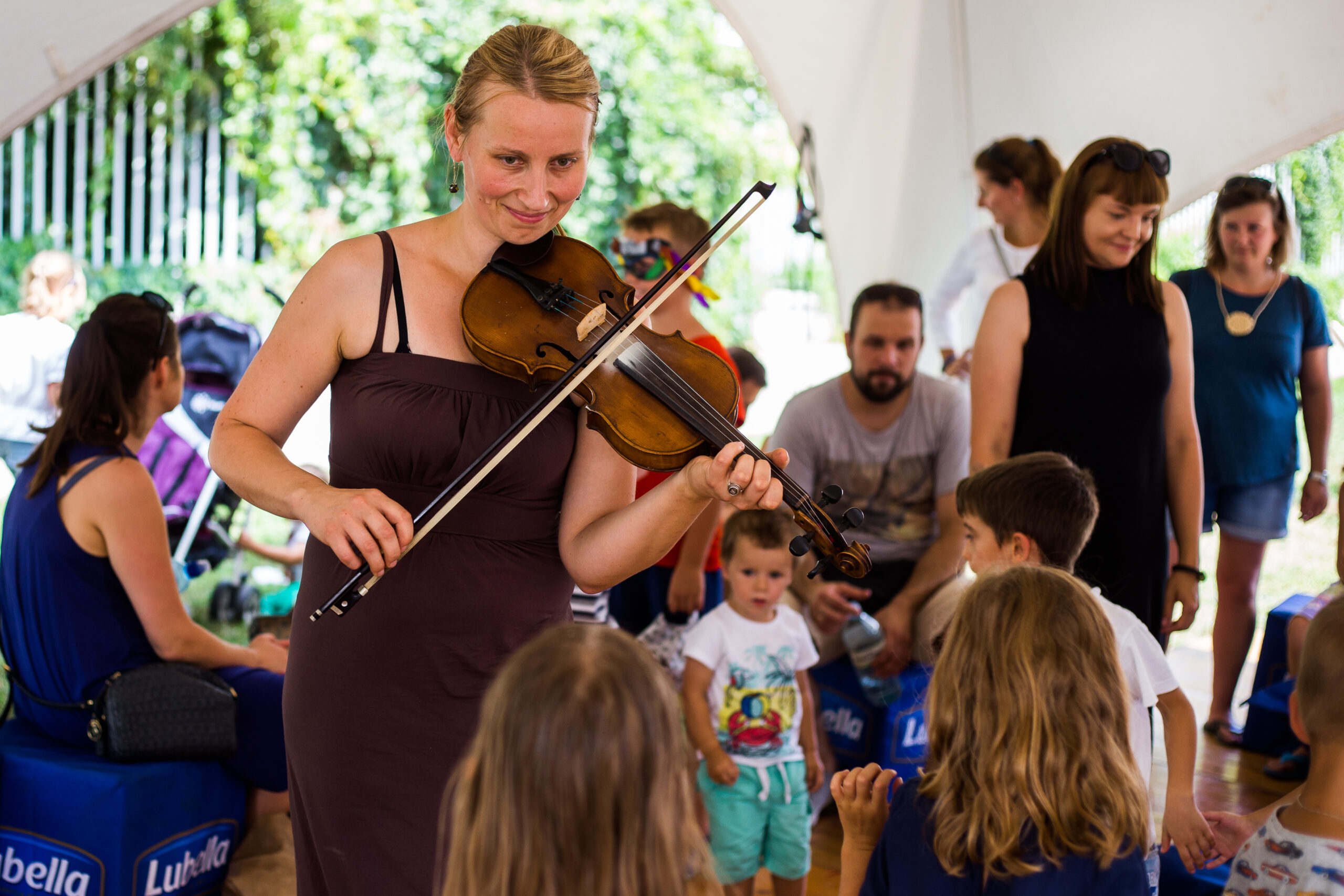 Katarzyna Zedel. Kobieta ubrana w długą brązową sukienkę na ramiączkach, gra na skrzypcach, uśmiecha się do zgromadzonych dzieci. W tle widzimy dzieci i rodziców, uczestników warsztatów.