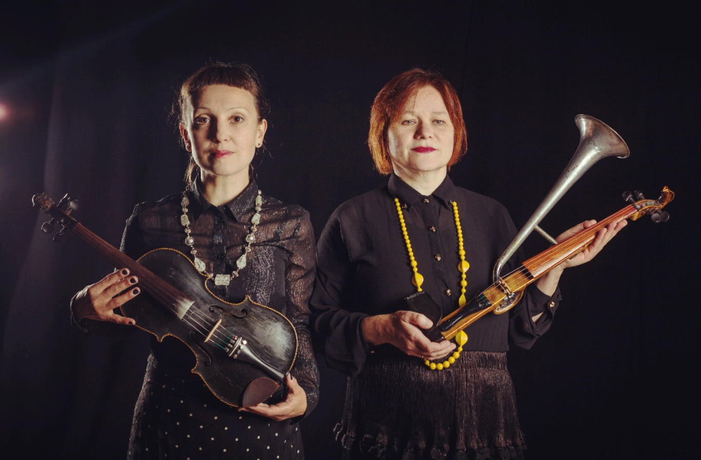 Na czarnym tle pozują dwie artystki, Katarzyna Szurman i Agata Harz. Kobiety mają ciemne ubrania i pomalowane na różowo usta. W rękach trzymają instrumenty - skrzypce i skrzypotrąbę.