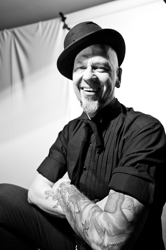 Czarno-biały portret Ofala Deriglasoffa, muzyka. Uśmiechnięty mężczyzna w czarnej koszuli i przekrzywionym kapeluszu na głowie. Jego ręce pokrywają tatuaże.