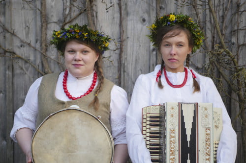 Na tle drewnianej ściany pozują dwie członkinie kapeli Kanka Franka. Kobiety ubrane w jasne stroje, czerwone korale i wianki uplecione z żywych kwiatów, trzymają w rękach instrumenty - bęben obręczowy i harmonię.