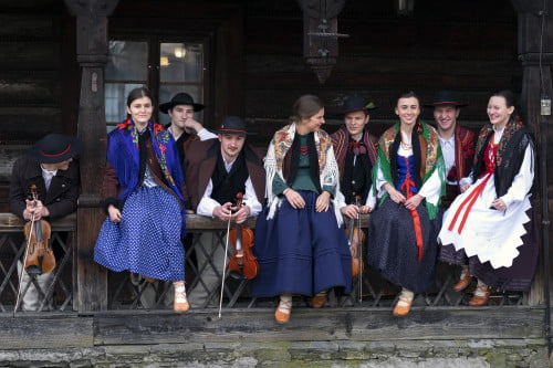 Kapela Mała Ziemia Suska w strojach regionalnych na tle starego drewnianego budynku. Kobiety siedzą z przodu na poręczy, za nimi stoją mężczyźni trzymający w rękach skrzypce.