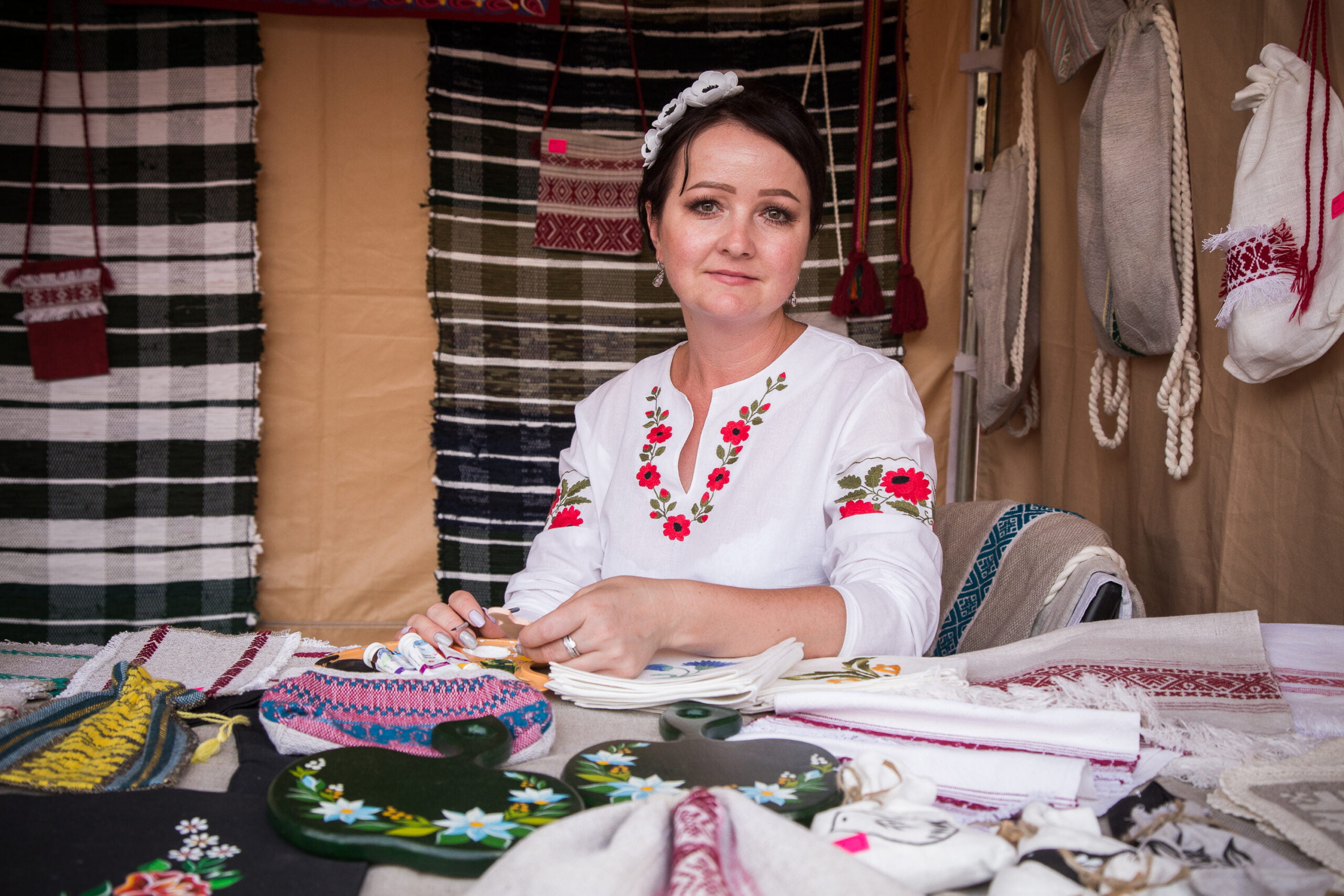 [Młoda kobieta w ludowym stroju, wokół wyroby ludowe. Haftowane tkaniny i inne.]