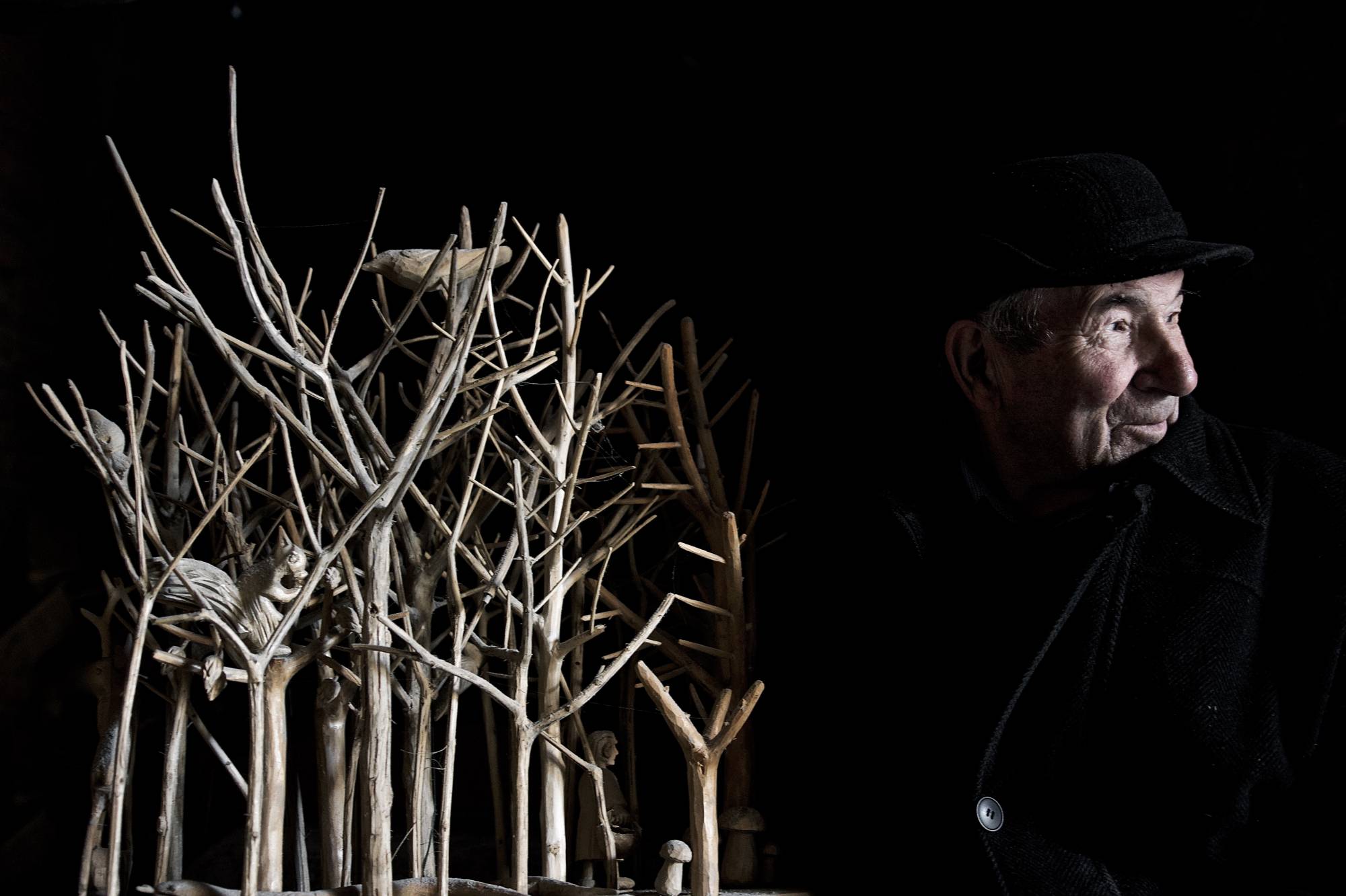 Starszy mężczyzna ubrany w czarny płaszcz stoi koło sztucznych drzew. Otacza go czarne tło.