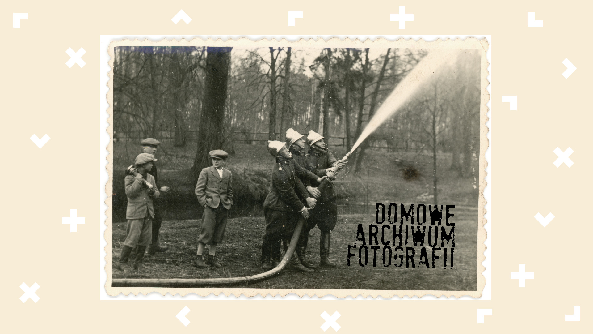 Czarno-biała fotografia strażaków z początku XX wieku podczas gaszenia pożaru. Przyglądają im się młodzi mężczyźni.