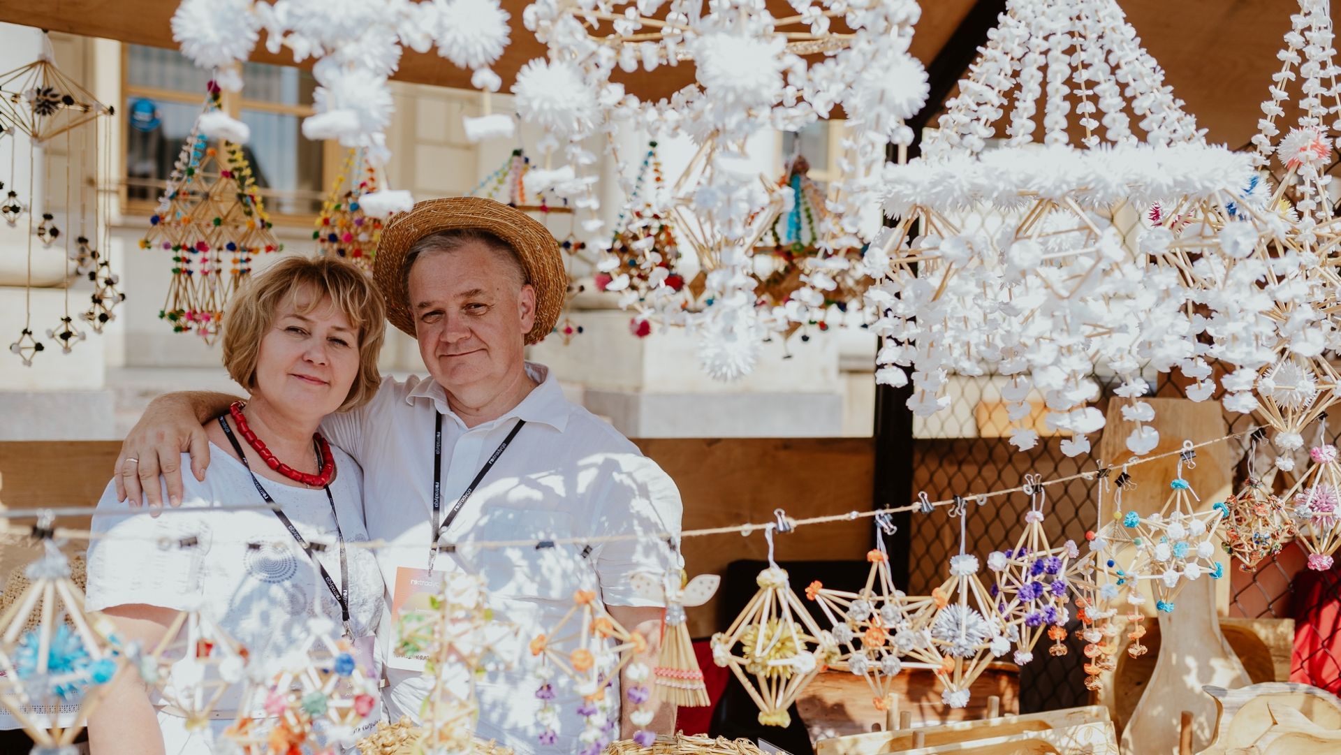 Kobieta i mężczyzna stoją przytuleni na stoisku. Sprzedają na nim pająki ludowe ze słomy.