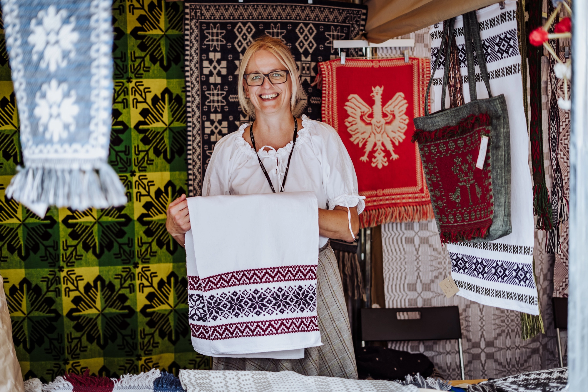 Portret twórczyni. Kobieta w białej bluzce stoi na tle swojego stoiska. W rękach trzyma jasną, utkaną przez siebie tkaninę.