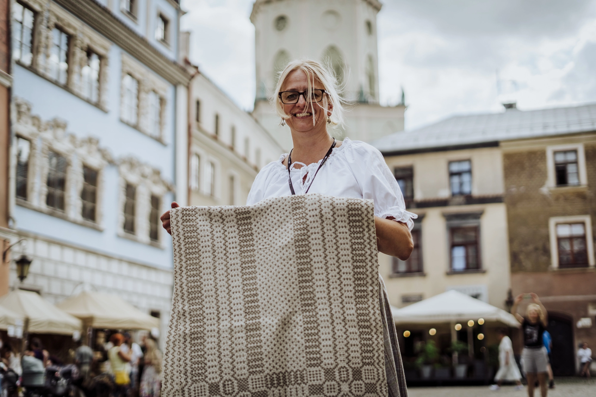 Portret twórczyni. Kobieta w białej bluzce stoi na tle Wieży Trynitarskiej i sąsiadujących z nią kamienic. W rękach trzyma jasną, utkaną przez siebie tkaninę.