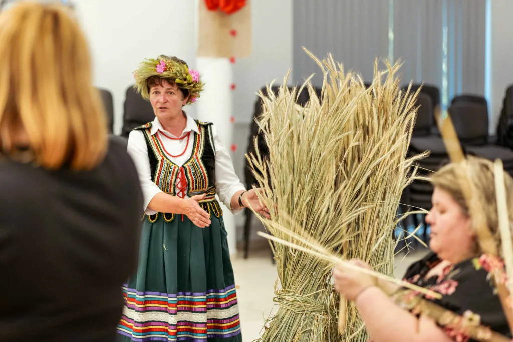 Zdjęcie. Instruktorka w ludowym stroju stoi obok dużego snopu siana. Mówi do uczestników warsztatów.