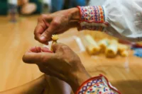 Zdjęcie. Dłonie kobiety w stroju ludowym i dziecka. Artystka pomaga dziecku zawiązać węzeł na słomie.
