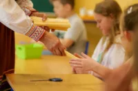 Zdjęcie. Kobieta w ludowym stroju rozdaje dzieciom wiązki słomy. Dłonie dziecka biorą od niej wiązkę.