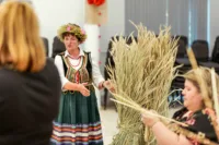 Zdjęcie. Instruktorka w ludowym stroju stoi obok dużego snopu siana. Mówi do uczestników warsztatów.