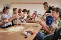 Zdjęcie. Skupione kobiety przy stole. Zaplatają słomę w grube warkocze.