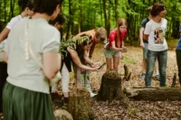 Zdjęcie. Las. Uczestnicy pochylają się nad pniakiem drzewa, aby zrobić zdjęcie.