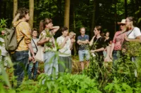 Zdjęcie. Las. Grupa stoi wokół przewodniczki i tłumaczki, które rozmawiają z jedną z uczestniczek.