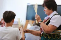 Zdjęcie. Chłopiec pracuje nad słomianą kukiełką. Pomaga mu stojąca obok prowadząca.