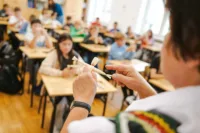 Zdjęcie. Szkolna sala. Dzieci siedzą w ławkach. Prowadząca trzyma w dłoni laleczkę i wyjaśnia, jak ją wykonać.