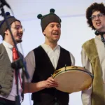 Trzech mężczyzn w jasnych koszulach, kamizelkach i czapkach. Śpiewają kolędy.