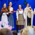 Trzy kobiety w strojach ludowych i dwóch mężczyzn. Jeden w stroju kozy. Śpiewają. Obok siedzi muzyk z suką biłgorajską.