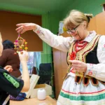 Twórczyni w stroju ludowym pokazuje dziecku gotowego pająka ze słomy i bibuły.