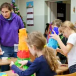 Prowadząca pokazuje dzieciom, jak oklejać bibułą kartonową czapę.