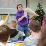 Prowadząca pokazuje dzieciom, jak dociąć karton na czapkę.