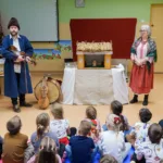 Sala pełna dzieci. Przed nimi prowadzący zajęcia i mała drewniana szopka z figurkami.