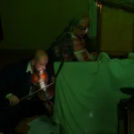 Ciemna sala. Artystka siedzi za szopką i porusza umieszczonymi w niej lalkami. Obok na ziemi siedzi artysta, który gra na skrzypcach.