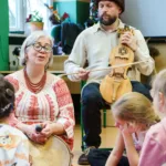 Artyści w strojach ludowych śpiewają z dziećmi. Mężczyzna gra na instrumencie. To suka biłgorajska.