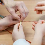 Dłonie dorosłej pomagają dziecku zawiązać węzeł na słomie.