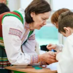 Twórczyni w stroju ludowym pochyla się nad dzieckiem, żeby pomóc mu zrobić aniołka ze słomy.